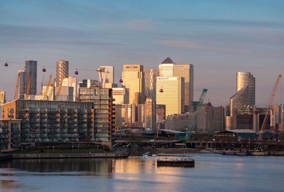 A daytime view of the Canary Wharf skyline in London, featuring several modern high-rise office buildings with glass facades, some of which are decorated with company logos such as Barclays. In the foreground, a construction site includes cranes, one green and one yellow, actively engaged in lifting and positioning building materials. To the lower left, a residential building with multiple balconies is situated near the riverbank, with a few small boats visible on the water. The scene shows a typical urban landscape associated with a busy business district, highlighting the ongoing building and construction activities that are part of the area's development. The sky is partly cloudy with soft lighting, and the river reflects the structures and cranes, emphasizing the active loading process relevant to house and furniture removals services in the Docklands area, as covered by Man With Van Docklands.