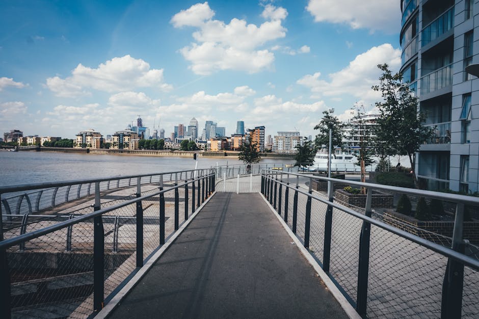 A wide pedestrian ramp with a metal railing on both sides leading down to a waterfront area in Docklands, Canary Wharf. The ramp has a smooth, dark surface with visible shadows cast by the railing. To the right, there is a modern, curved residential building with balconies and large windows, partially shaded by a few trees. On the left, the river stretches across the scene, with a view of London's skyline in the distance, featuring various tall office buildings and cranes under a partly cloudy sky. The environment appears well-maintained, with clean walkways, outdoor lighting fixtures, and some landscaping elements. This setting exemplifies an accessible pathway often involved in urban home relocation or furniture transport logistics, with the natural atmosphere and infrastructure supporting efficient loading, unloading, and moving activities, as highlighted by Man With Van Docklands' comprehensive removals and moving services.