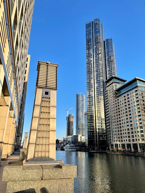 A daytime view of the Canary Wharf skyline in London, featuring several modern high-rise office buildings with glass facades, some of which are decorated with company logos such as Barclays. In the foreground, a construction site includes cranes, one green and one yellow, actively engaged in lifting and positioning building materials. To the lower left, a residential building with multiple balconies is situated near the riverbank, with a few small boats visible on the water. The scene shows a typical urban landscape associated with a busy business district, highlighting the ongoing building and construction activities that are part of the area's development. The sky is partly cloudy with soft lighting, and the river reflects the structures and cranes, emphasizing the active loading process relevant to house and furniture removals services in the Docklands area, as covered by Man With Van Docklands.