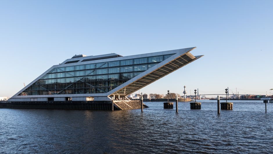 A modern, angular building with multiple sloped glass and metal roofs, situated on the edge of a calm river during sunset. The reflection of the building is visible in the water, which appears smooth 