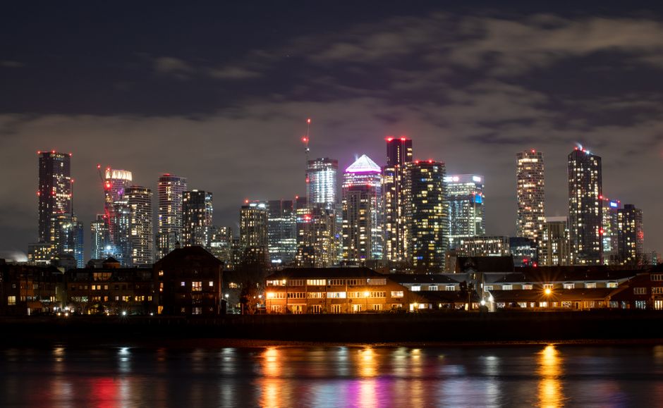Night-time view of the London skyline in the Docklands area, with illuminated high-rise office buildings and residential skyscrapers, some with red aviation warning lights on their rooftops. In the foreground, low-rise residential buildings with lit windows sit along the riverbank, reflecting the city lights on the water's surface. The sky is partly cloudy, adding contrast to the brightly lit structures. This image captures the urban landscape that relocations or moving services, such as those provided by Man With Van Docklands, might involve when planning furniture transport or home relocation in the Isle of Dogs and Millharbour area.