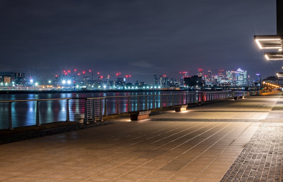 Night-time view of the London skyline in the Docklands area, with illuminated high-rise office buildings and residential skyscrapers, some with red aviation warning lights on their rooftops. In the foreground, low-rise residential buildings with lit windows sit along the riverbank, reflecting the city lights on the water's surface. The sky is partly cloudy, adding contrast to the brightly lit structures. This image captures the urban landscape that relocations or moving services, such as those provided by Man With Van Docklands, might involve when planning furniture transport or home relocation in the Isle of Dogs and Millharbour area.