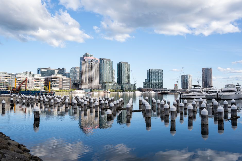 A wooden dock extending into a calm body of water in the Docklands area, with several green and black mooring posts along the edges. In the background, there is a modern city skyline featuring high-ri